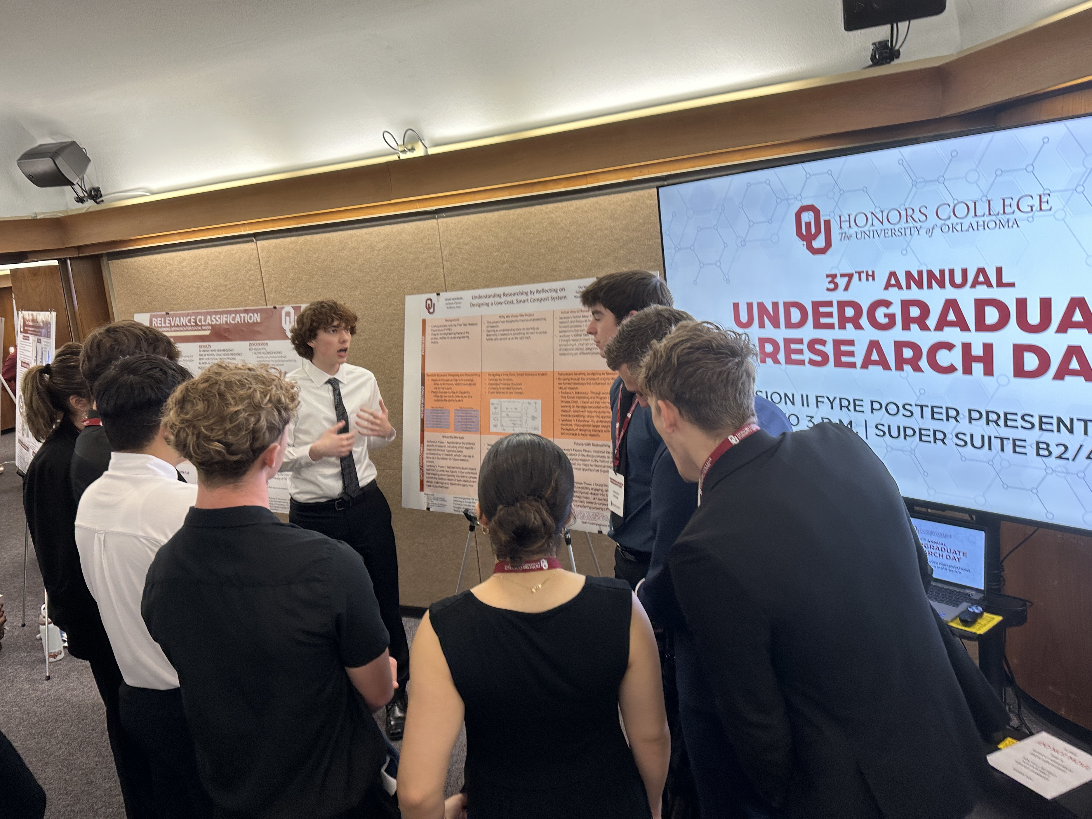 Anthony Voci speaking to the left of the poster with Jackson Clymer standing to the right, surrounded by interested peers.